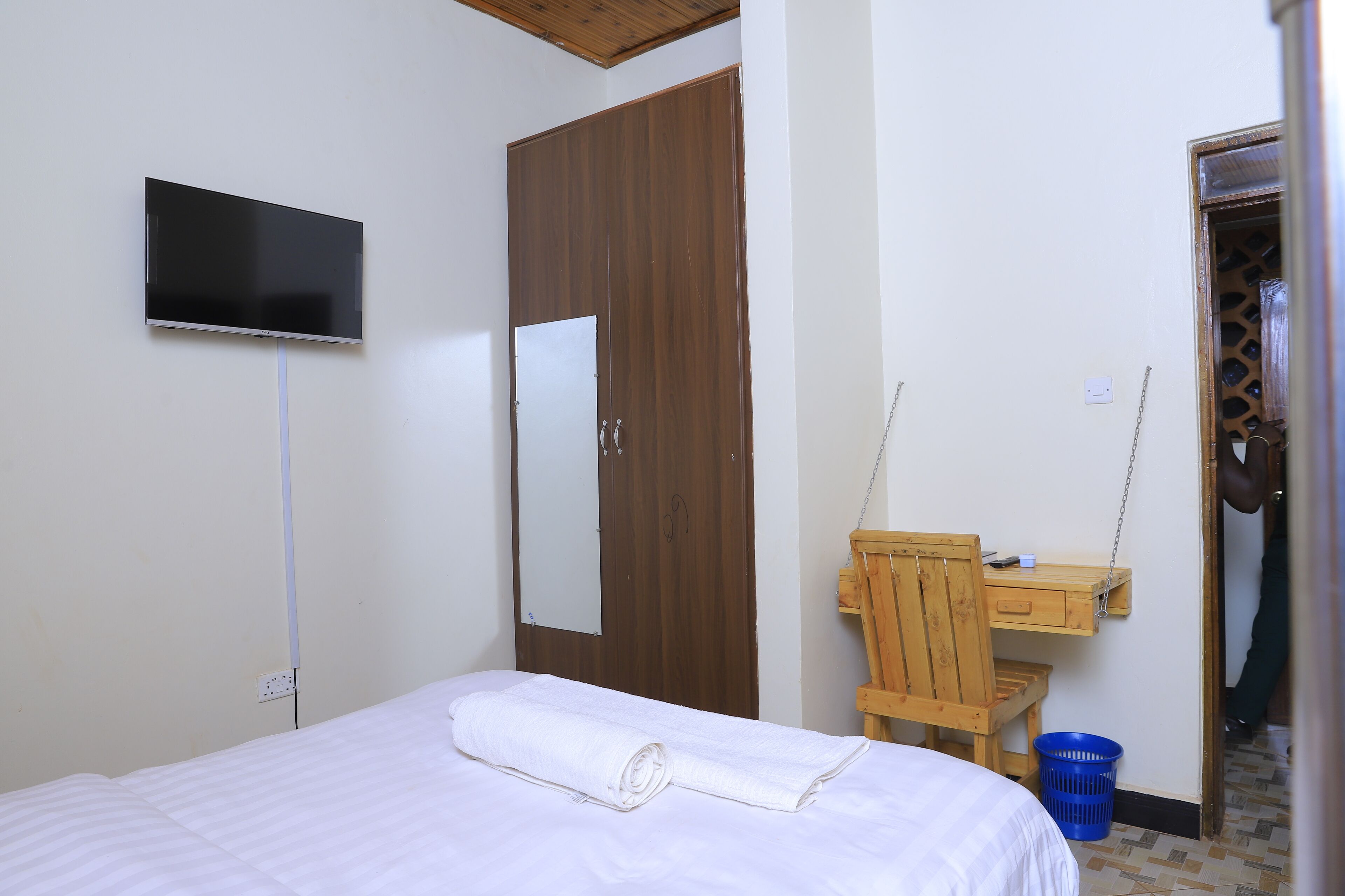 A hotel room with a bed, a TV mounted on the wall, a wooden wardrobe, a desk with a chair, and a blue laundry basket.