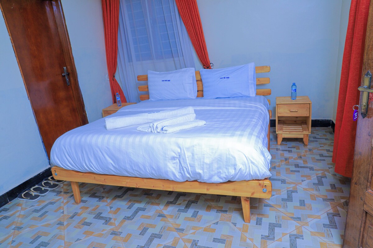 Una cama bien hecha con sábanas y almohadas blancas, una mesita de noche de madera con una botella de agua y un suelo estampado.