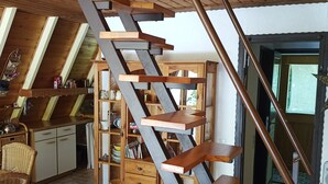 Una escalera espiral de madera en una habitación estilo loft con techo alto, estanterías empotradas de madera y una zona de cocina con armarios y una mesa de comedor.
