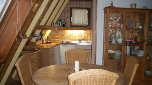 Un comedor con una mesa redonda y cuatro sillas de mimbre, un mueble de madera con puertas de vidrio y una cocina al fondo.
