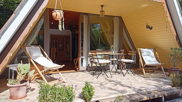 Un espacio exterior acogedor y de forma triangular con una terraza de madera, una mesa de comedor con sillas y una jardinera colgante.
