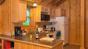 Une cuisine rustique aménagée dans une cabane en bois, avec un îlot central en bois, un réfrigérateur, un four à micro-ondes, une cuisinière et une fenêtre donnant sur un paysage verdoyant.