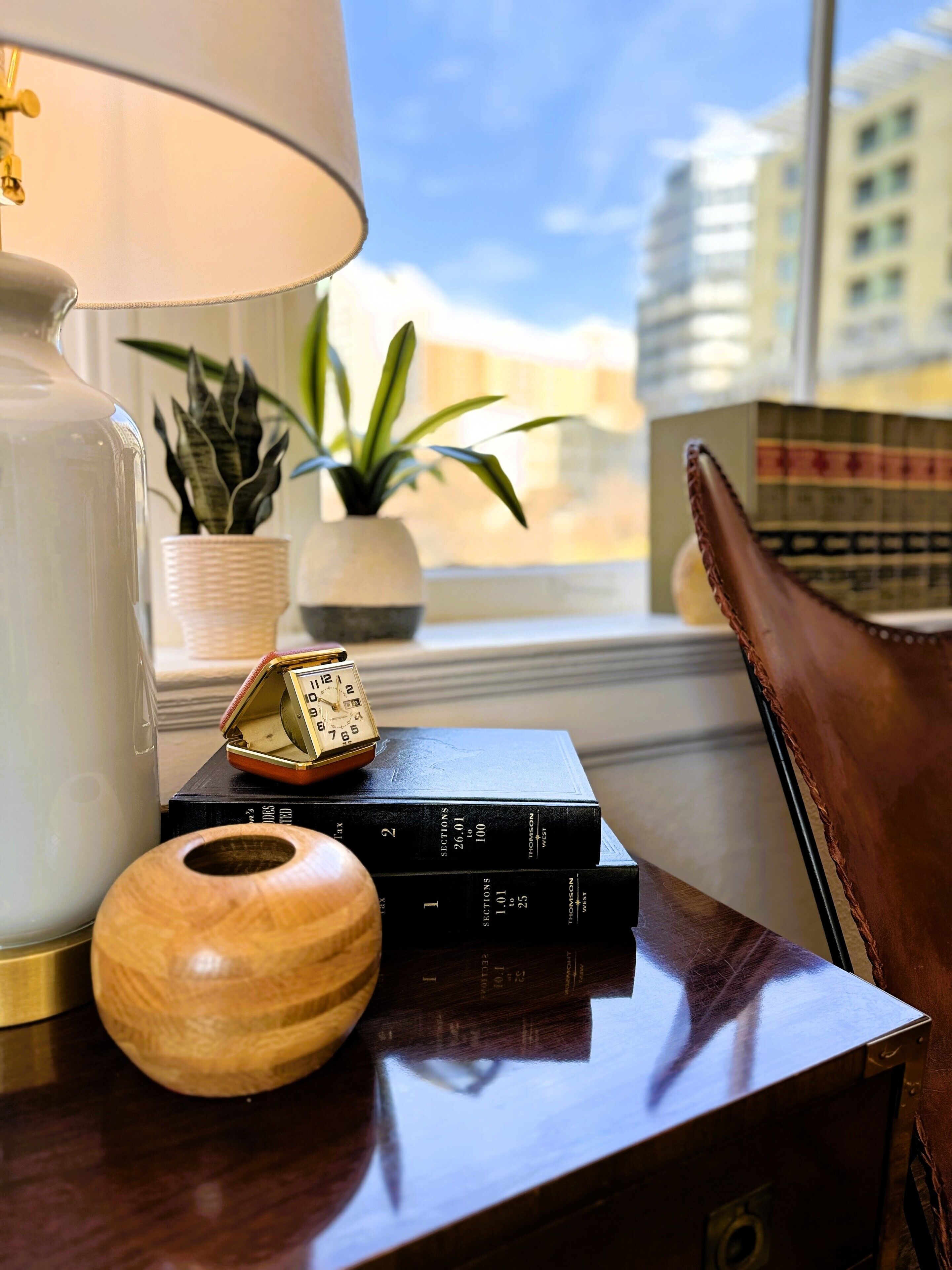 A wooden side table with a lamp, a wooden bowl, a stack of books, a small plant, and a leather chair.