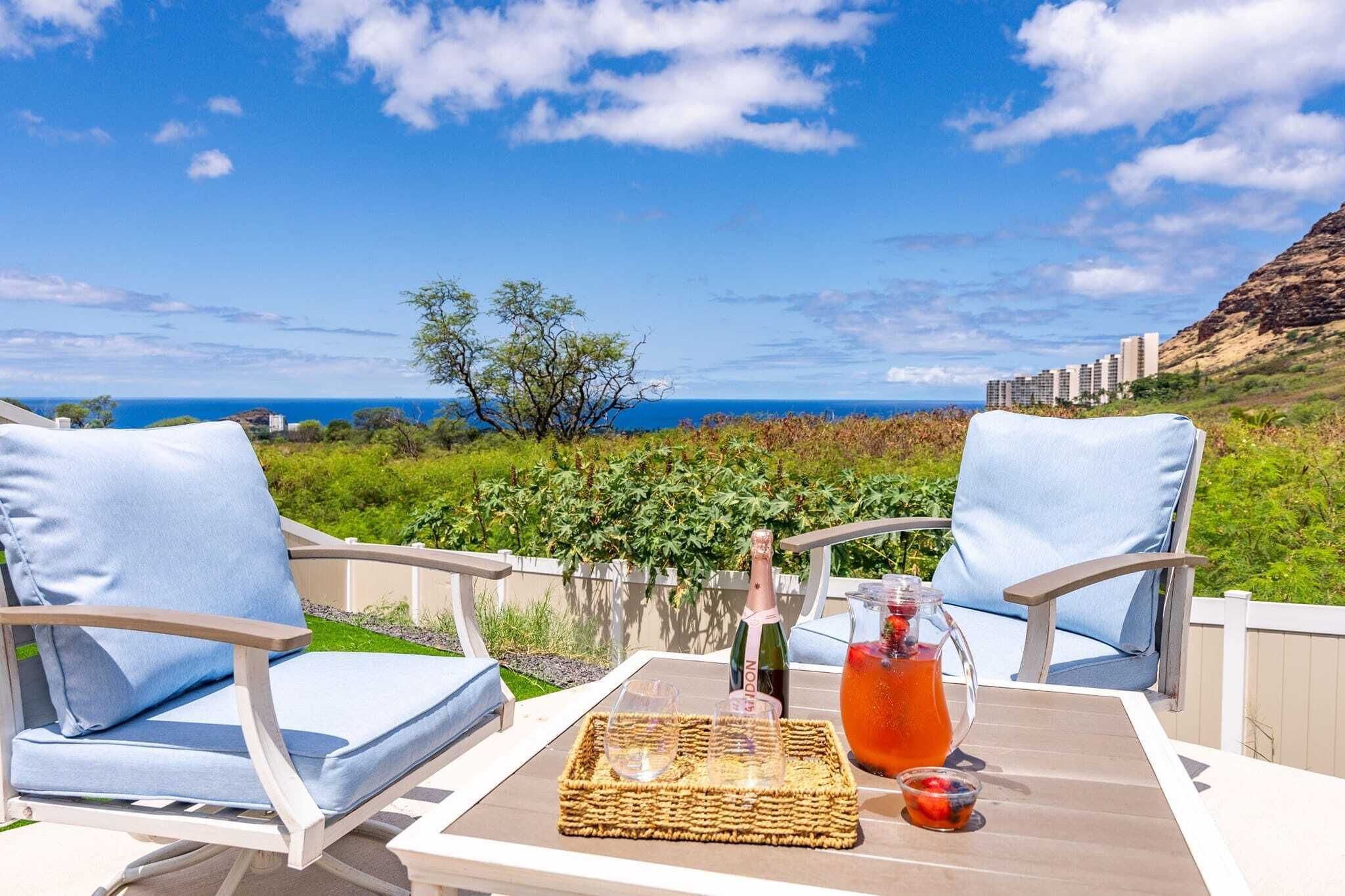 A terrace with two light blue cushioned chairs, a wicker tray with a bottle of champagne, glasses, and a pitcher of fruit drink.