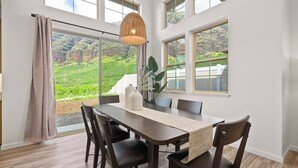 Ein Essbereich mit Tisch, Stühlen, einer Pendelleuchte und großen Fenstern mit Blick auf eine grüne Landschaft.