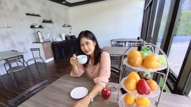 Une personne est assise à une table devant une assiette blanche et une tasse. En arrière-plan, on aperçoit un étal de fruits avec des oranges et des poivrons rouges, ainsi qu’un coin cuisine.