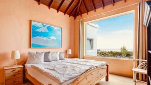 Ein Schlafzimmer mit einem hölzernen Bett, einem Nachttisch, einem Fenster mit Blick auf den Strand und einem Bild an der Wand.