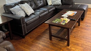 Un canapé d’angle en cuir marron foncé agrémenté de coussins décoratifs, une table basse en bois avec des magazines et une télécommande, et un plancher en bois.