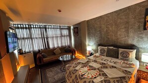 Ein Hotelzimmer mit Sofa, einem kleinen Tisch, einem Bett mit dekorativem Kopfteil, einem Fernseher und einem gemusterten Teppich.