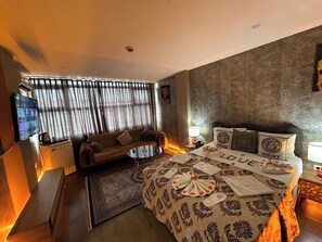 A hotel room with a sofa, a small table, a bed with a decorative headboard, a television, and a patterned rug. - Kral As Süite Hotel (Istanbul)