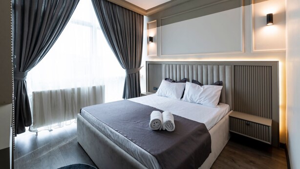 Ein Hotelzimmer mit einem großen Bett, einem Nachttisch, einer Kopfteil und einem fenster mit Vorhängen.