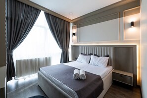 Una habitación de hotel con una cama grande, una mesita de noche, cabecero y una ventana con cortinas.