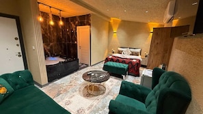 Una habitación de hotel moderna con suelo de mármol, una cama con colcha floral roja y negra, un sofá tapizado de color verde, una mesita redonda pequeña y un jacuzzi.
