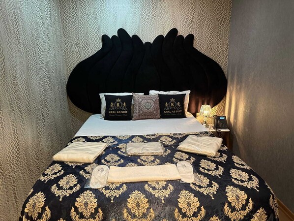 Una cama con cabecera negra, dos almohadas con el logotipo 'KRAL AS SUIT', una colcha estampada y toallas dobladas con cuidado.