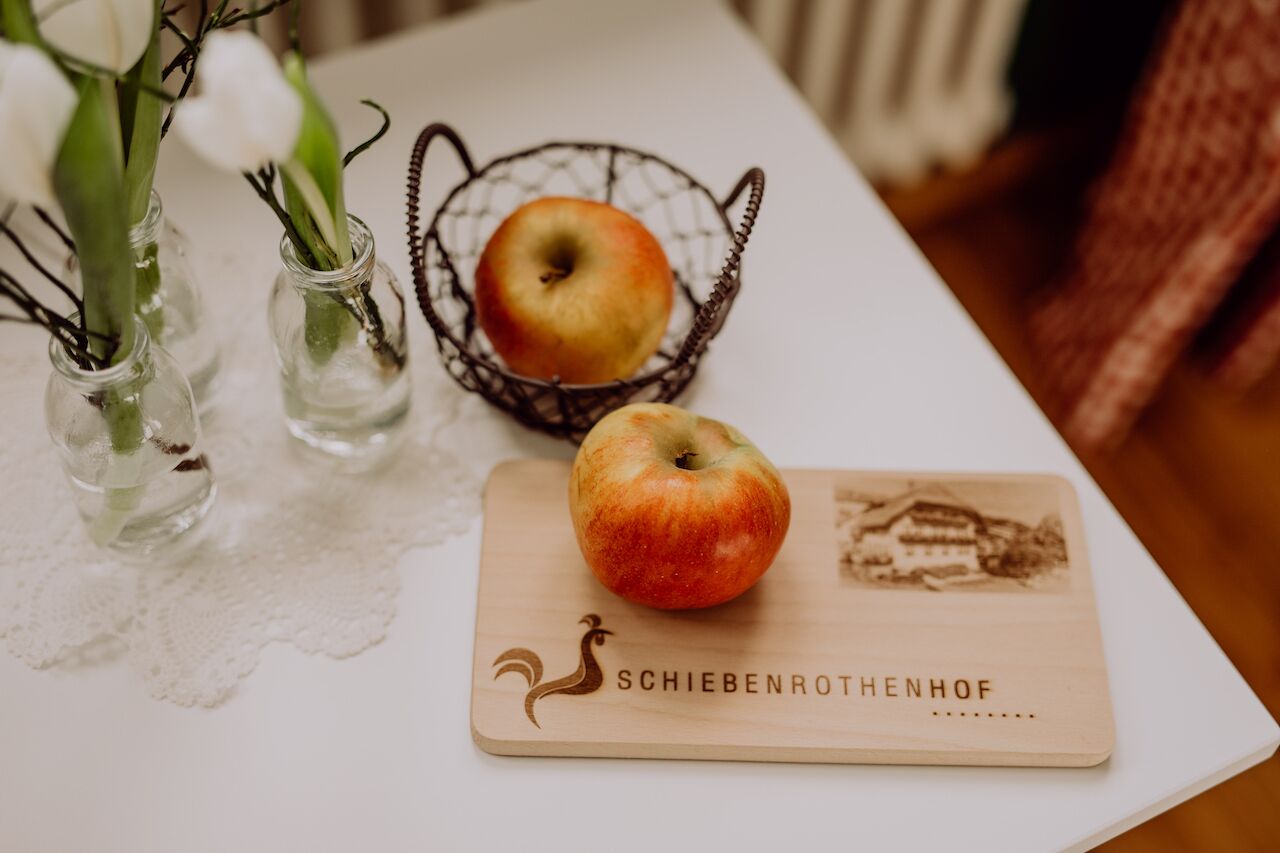 Une table avec une planche à découper en bois, un panier contenant une pomme et deux vases en verre avec des fleurs.