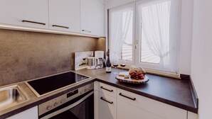 A modern kitchen with a built-in oven, a stovetop, and a sink. There is a window with sheer curtains, and a countertop with a cheese board and grapes. - Schiebenrothenhof (Simonswald)
