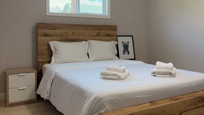 A bedroom with a wooden bed, white bedding, a nightstand, a framed picture, and a window with a view of the outside.