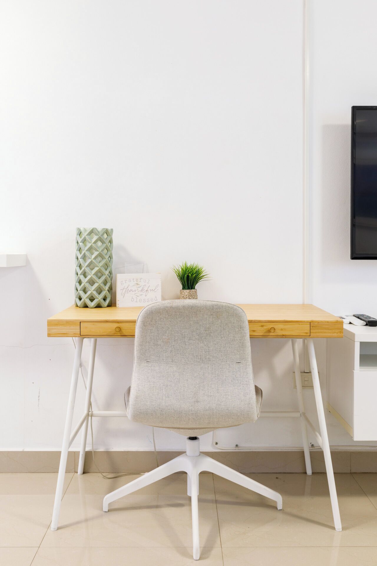 Un escritorio moderno con tapa de madera clara y patas blancas, una silla tapizada de gris con base de cinco brazos, una pequeña planta en maceta y un cartel con un mensaje positivo.