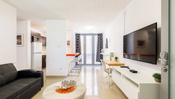 A modern living room with a black leather sofa, a round wicker coffee table, a flat-screen TV, and a kitchen area with a refrigerator and microwave. - Avila Suites (San Juan)