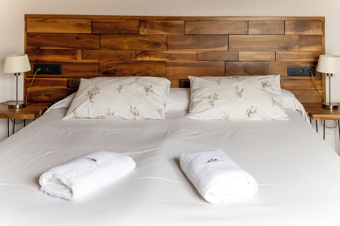 Una cama doble con cabecera de madera, dos almohadas con motivos florales, dos lámparas de noche y dos toallas enrolladas.