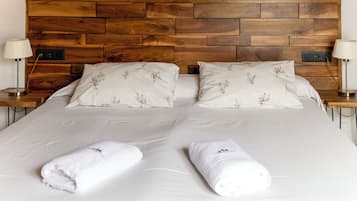 Una cama doble con cabecera de madera, dos almohadas con motivos florales, dos lámparas de noche y dos toallas enrolladas.