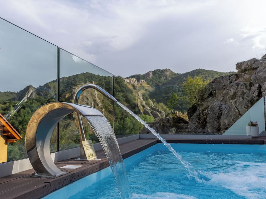 Piscina moderna al aire libre con caída de agua, rodeada por barandilla de vidrio y con vistas a un paisaje montañoso.