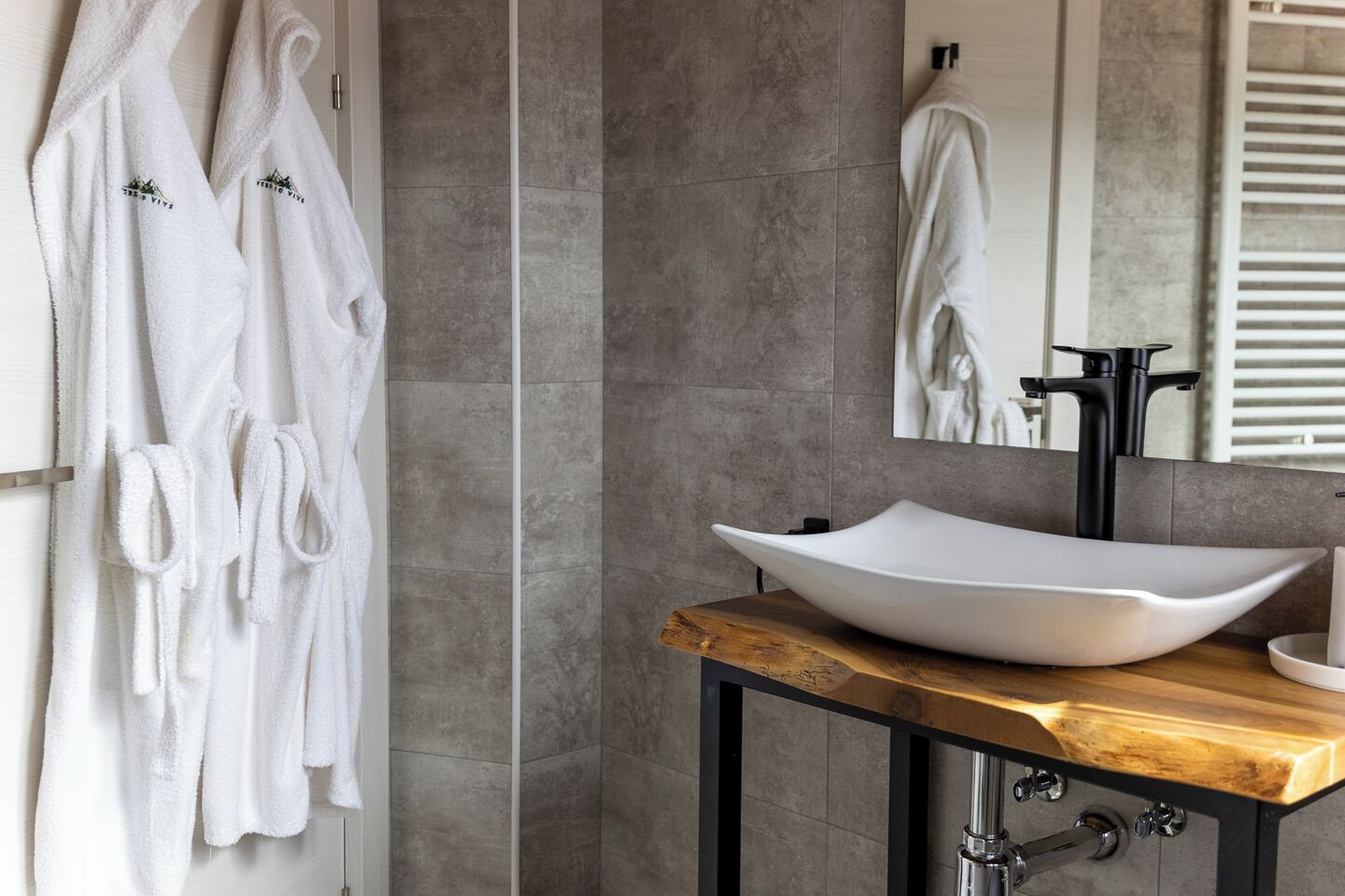 A bathroom with a large, white vessel sink on a wooden countertop, grey tiled walls, and two white towels hanging on a rack.