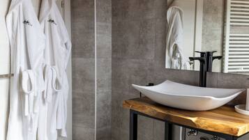 A bathroom with a large, white vessel sink on a wooden countertop, grey tiled walls, and two white towels hanging on a rack.