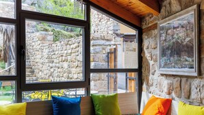 Ein gemütlicher Sitzbereich mit Steinwand und großen Fenstern, mit Blick auf ein Steinhaus und grüne Landschaft.