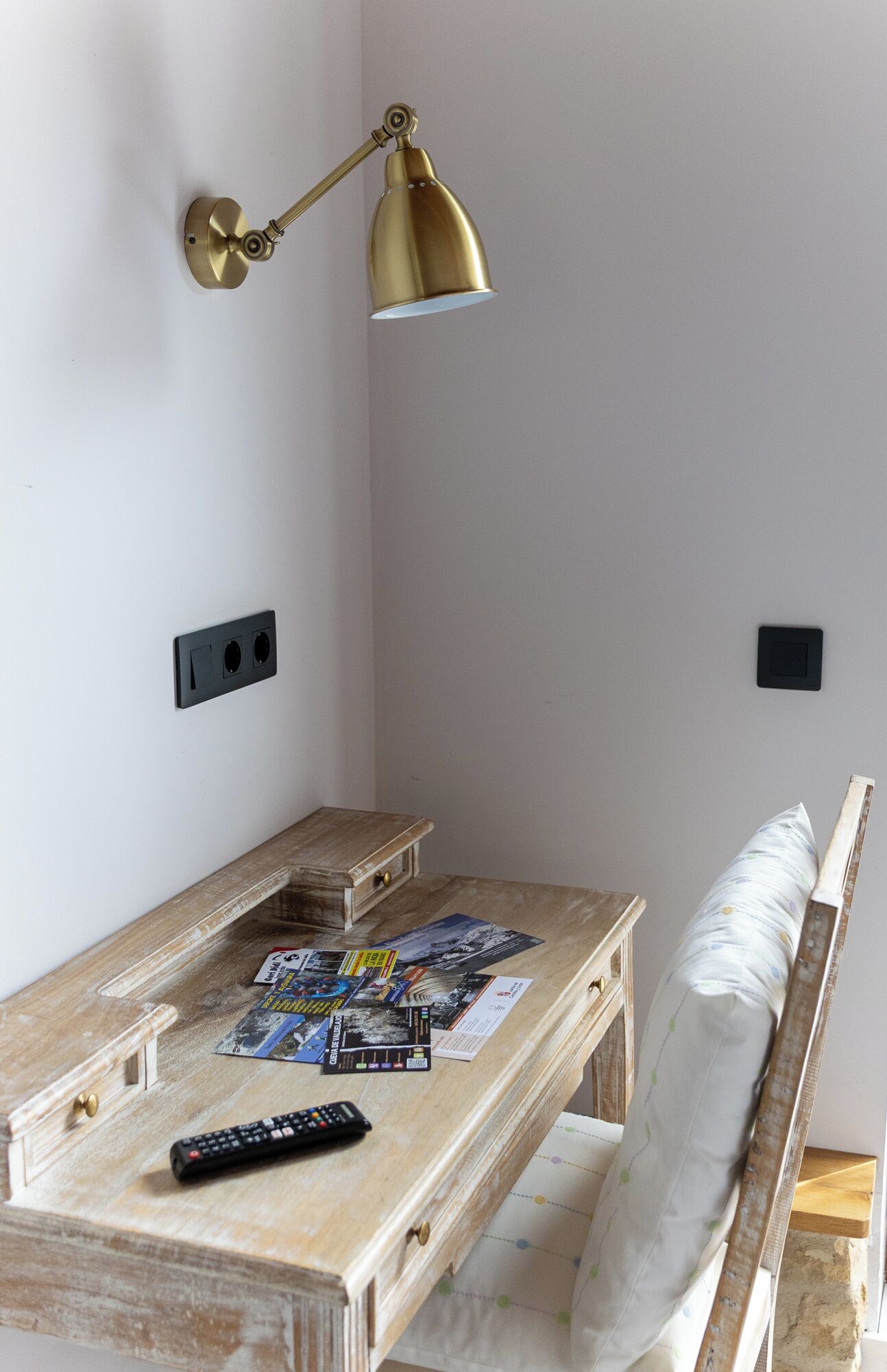 A small, cozy room with a wooden desk, a lamp, a remote control, and a bed with a pillow.