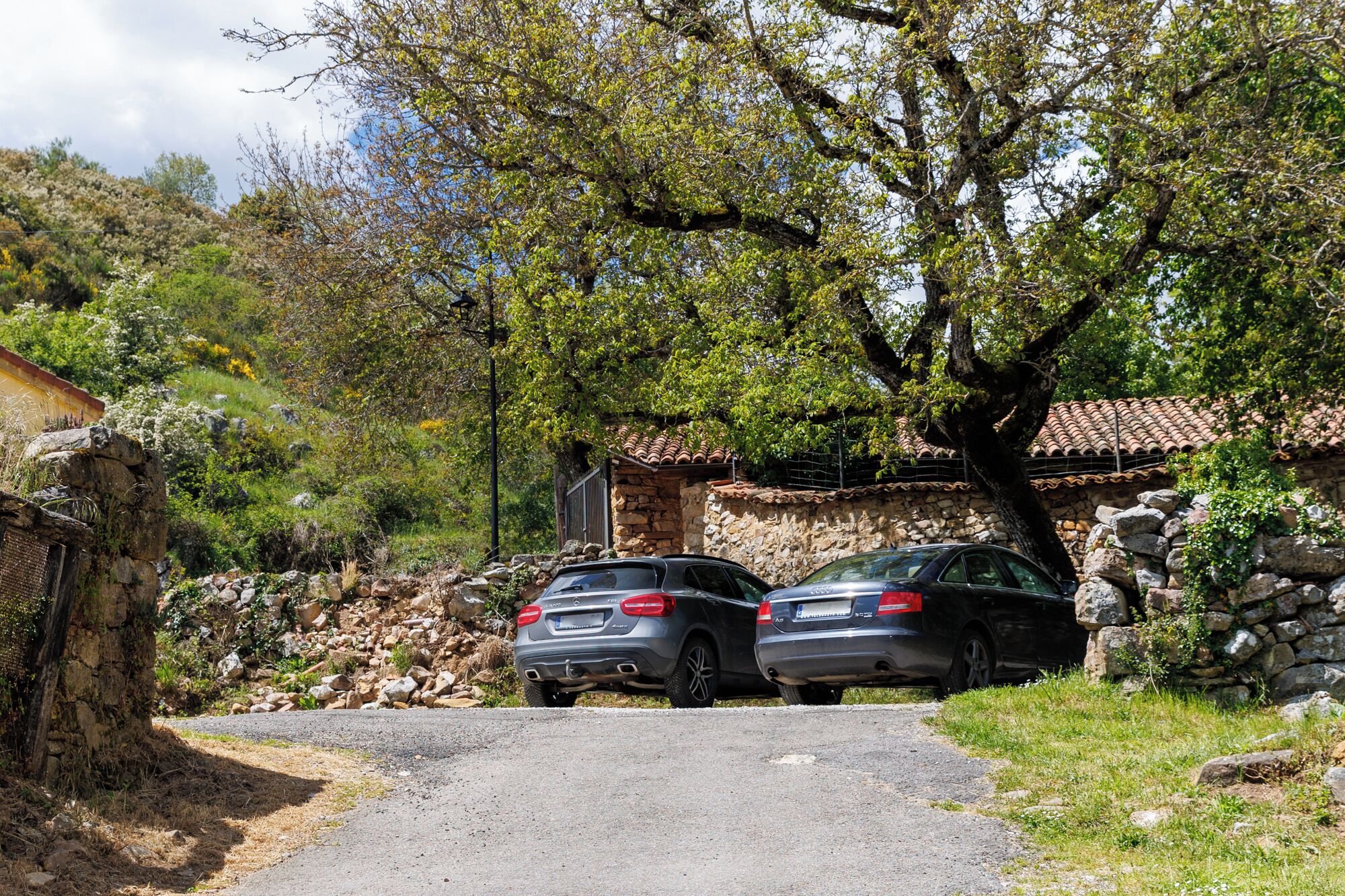 Deux voitures sont garées devant un bâtiment en pierre, surmonté d’un toit en tuiles, et entouré d’arbres et d’un mur de pierre.