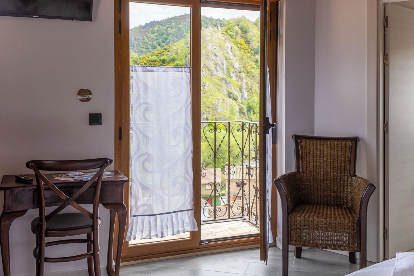 A room with a wooden desk, a wicker chair, and a balcony with a view of green hills.