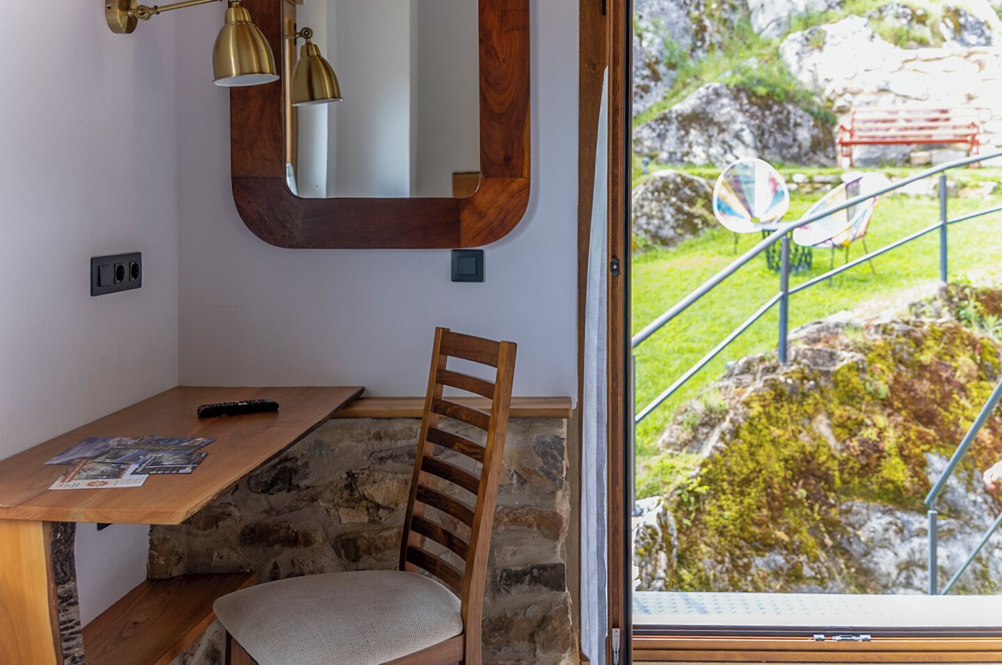 Une petite pièce avec un bureau en bois, une chaise, un miroir et une fenêtre donnant sur un jardin agrémenté d’une rambarde en métal.