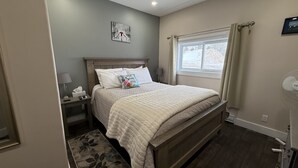 A bedroom with a bed, bedside tables, a window with curtains, a wall-mounted picture, and a floor lamp. - The Trailway Hub, ideally located in Deer Lake, 10 minutes from the airport (Deer Lake)