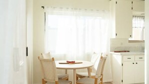 Un comedor con una mesa redonda y cuatro sillas, una cocina con armarios blancos y un ventanal con cortinas translúcidas.
