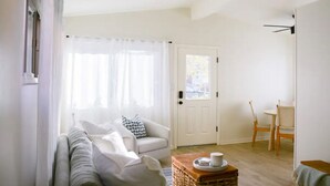 Una sala de estar luminosa con un sofá, una silla, una mesa con una taza y una puerta con un ventanal.