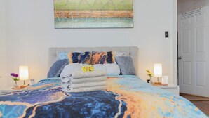 Ein ordentlich bezogenes Bett mit einer farbenfrohen Decke, ein Gemälde darüber und zwei Nachttische mit Blumen.