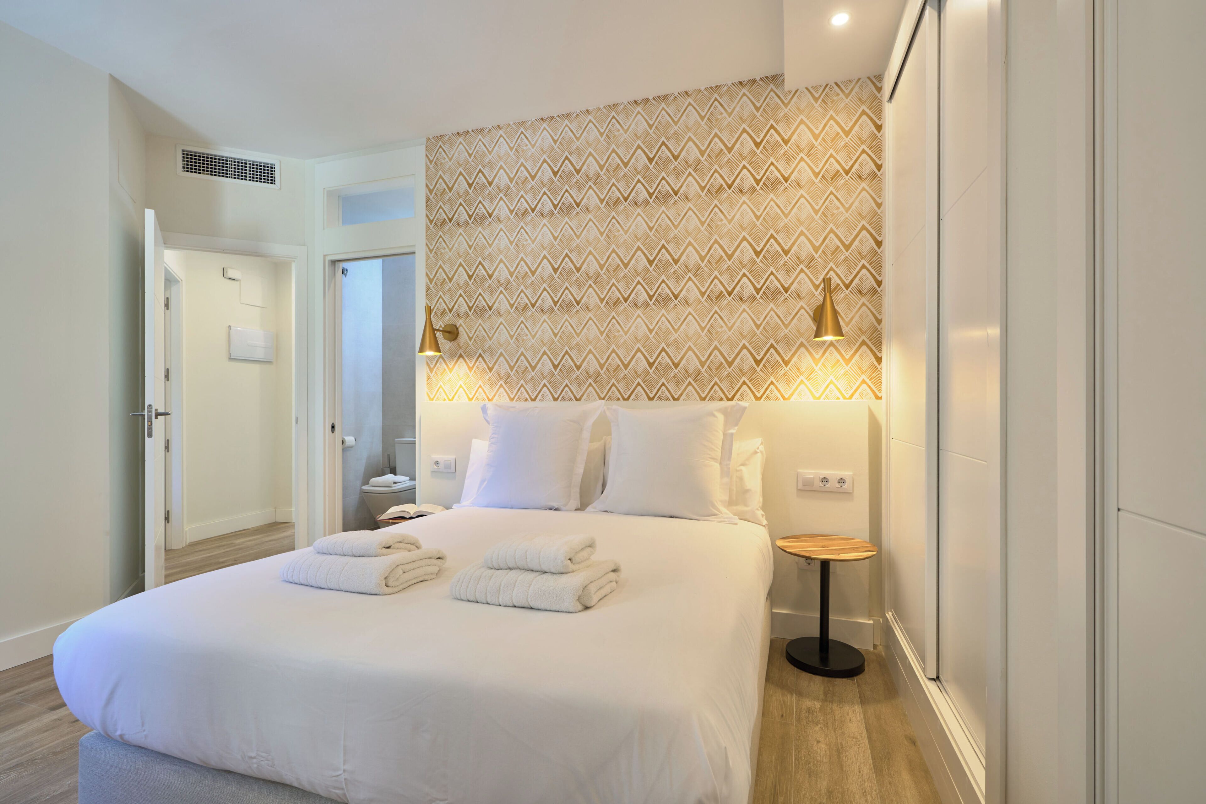 Ein modernes Hotelzimmer mit einem ordentlich bezogenen Bett, einem Nachttisch mit Lampe und einer gemusterten Wand dahinter.