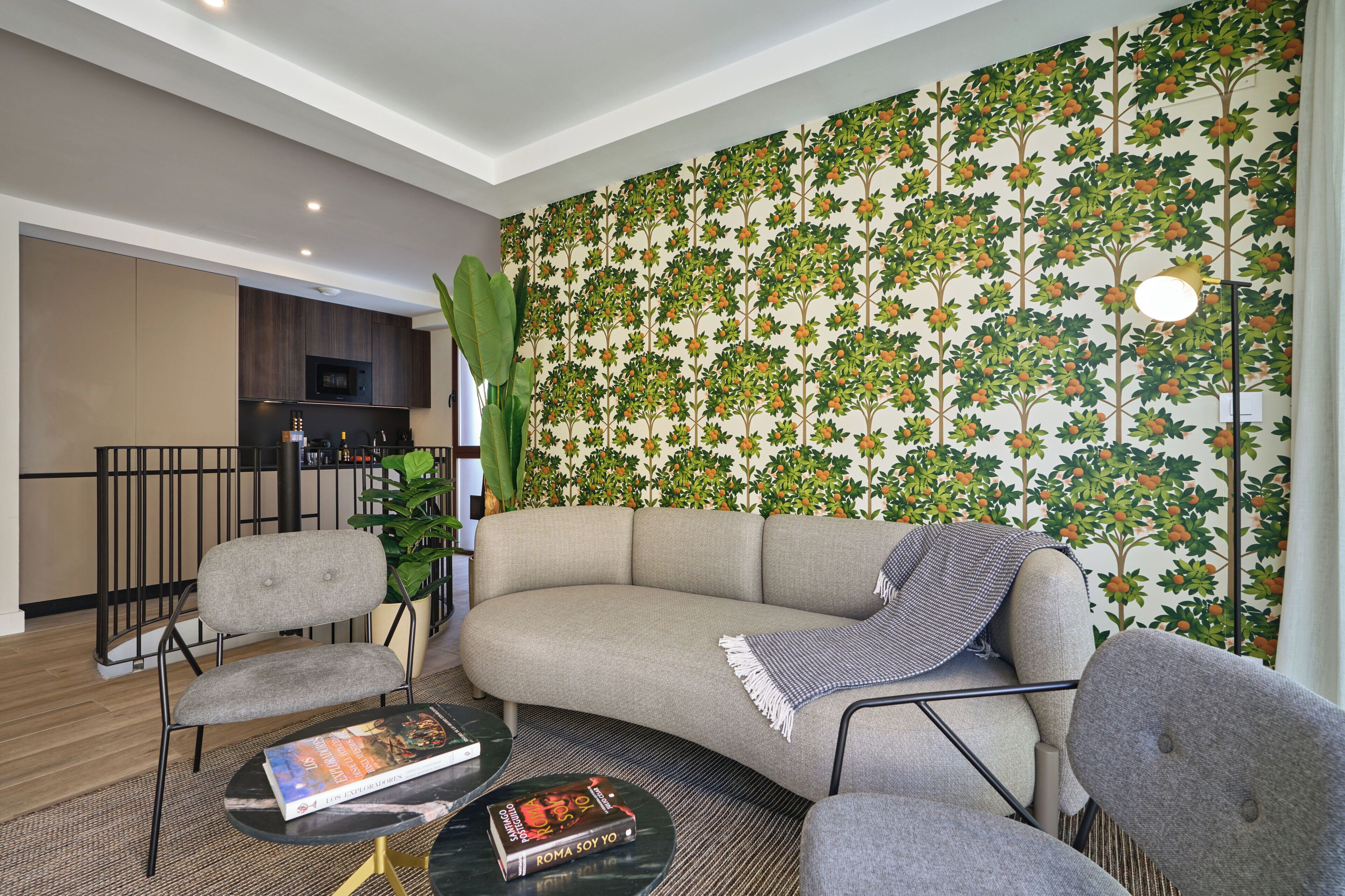 En moderne stue med en buet sofa, to lænestole, et sofabord og en reol. Der er også en stor potteplante og en væg med blomstermønster at se.
