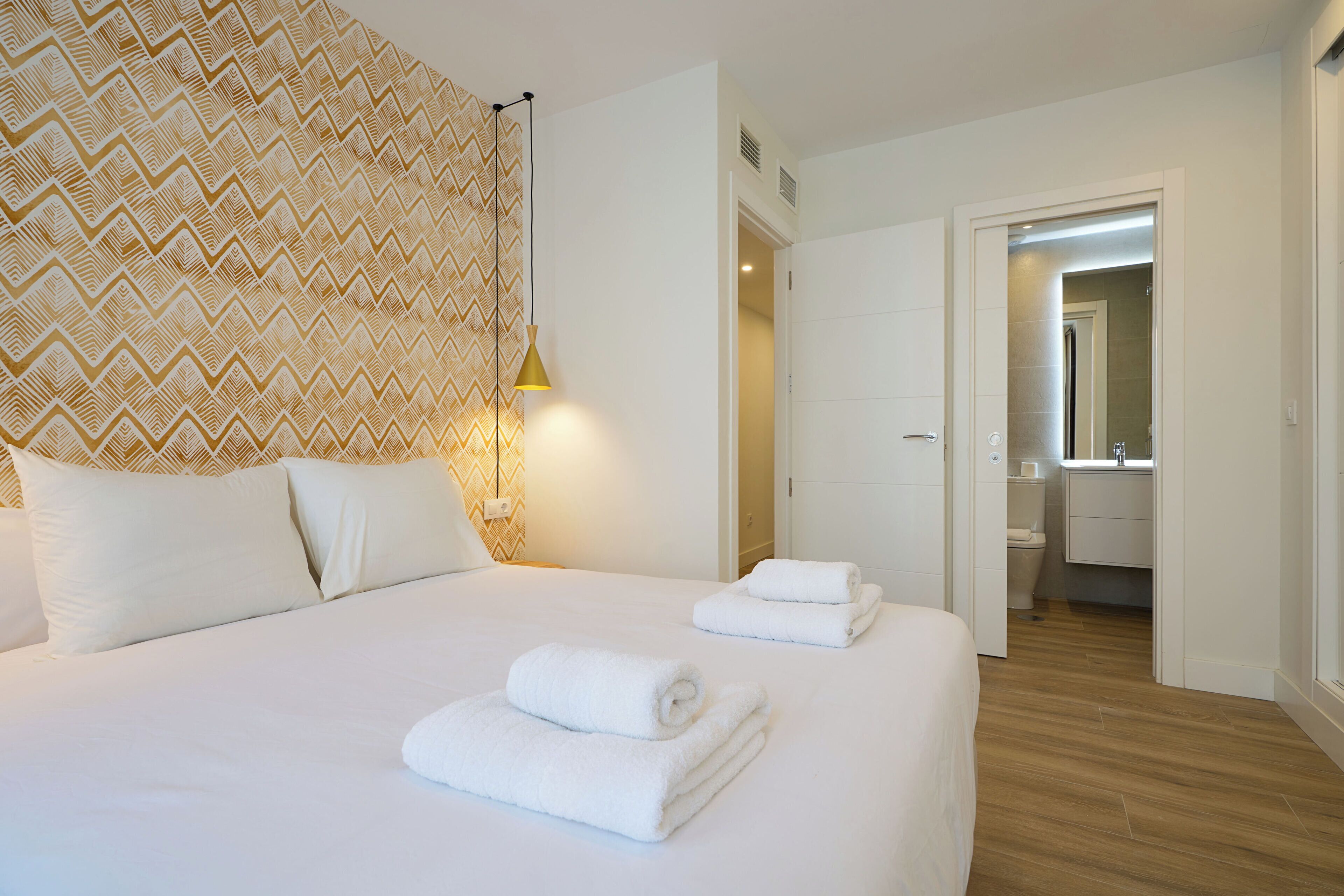 A hotel room with a large bed, two white towels, a patterned wall, and a bathroom visible through an open door.