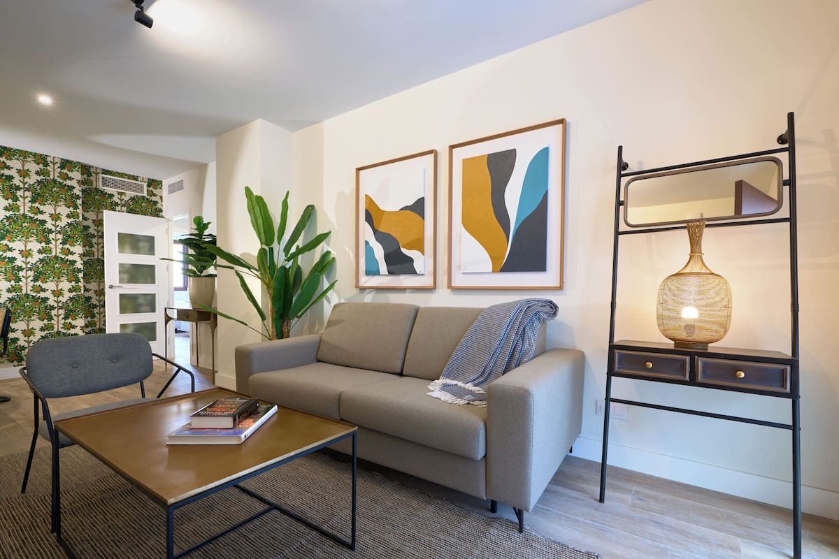 Un salon moderne avec un canapé, une table basse et une table d’appoint sur laquelle se trouve une lampe. Deux tableaux abstraits ornent le mur, et une plante en pot est également présente.