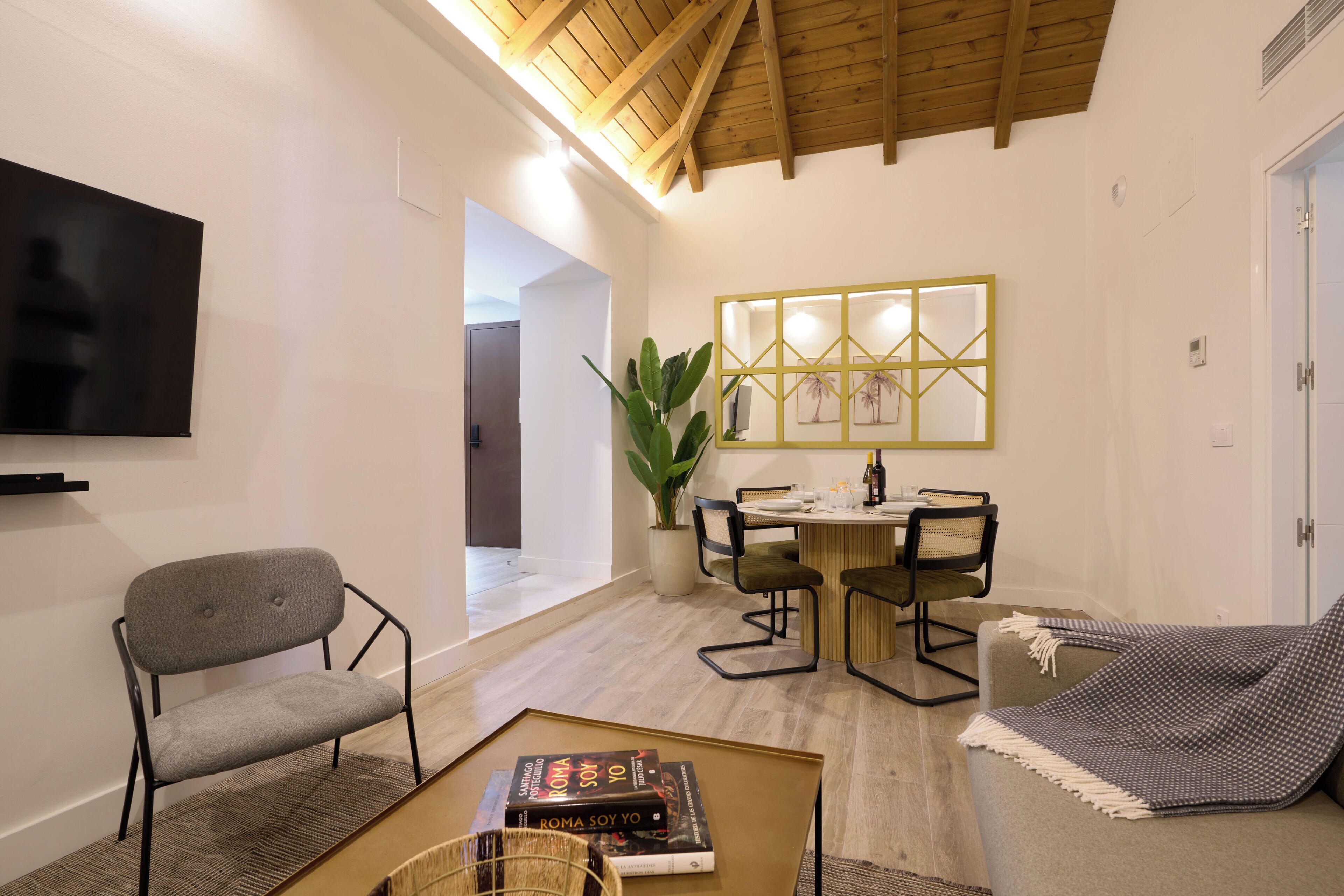 Una sala de estar moderna con techo de madera, televisor de pantalla plana, zona de comedor con mesa y sillas, y un rincón de descanso acogedor con una manta.