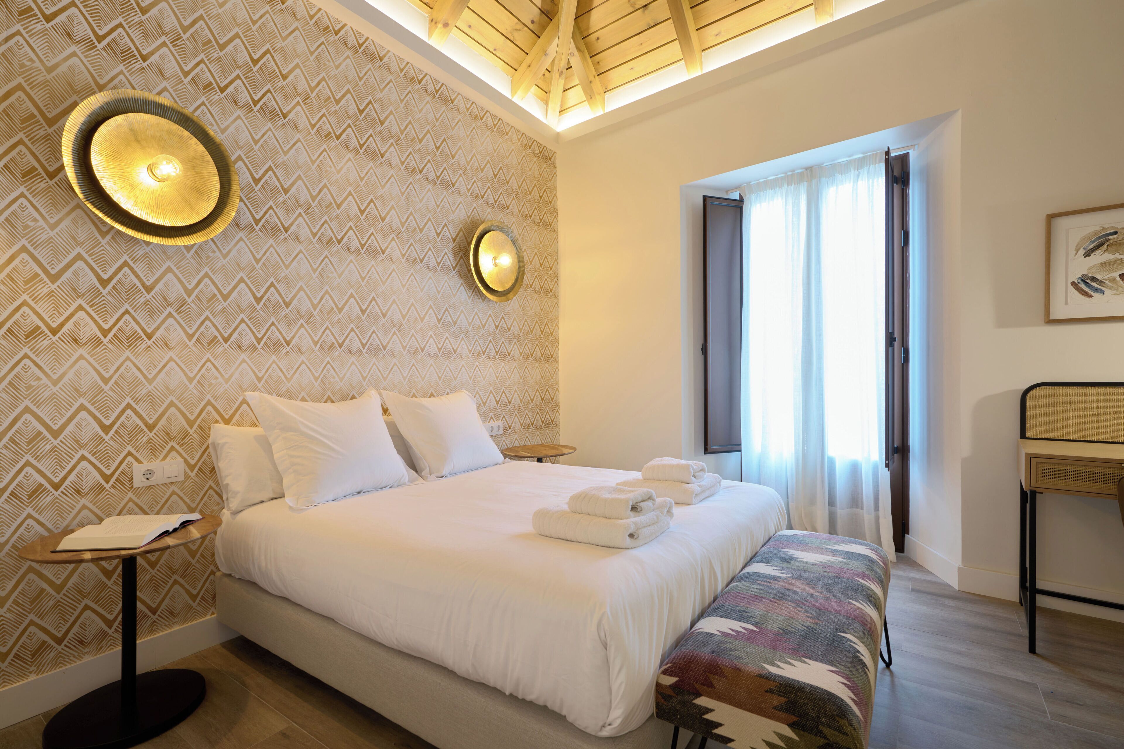 Une chambre d’hôtel avec un grand lit, une petite table de chevet, un banc, une horloge murale et une fenêtre avec des rideaux.