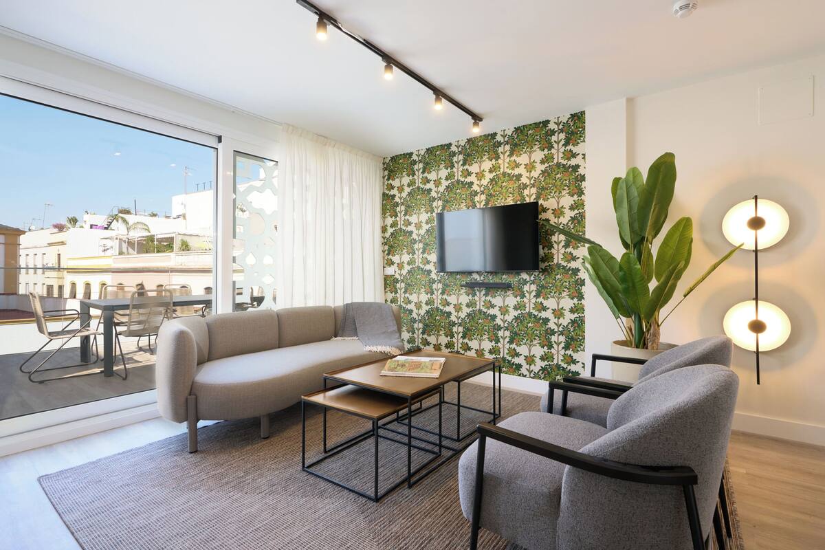 Un salon moderne comprenant un canapé, une table basse et des fauteuils, un téléviseur à écran plat fixé sur un mur recouvert d’un papier peint à motifs floraux, une grande plante et un coin repas avec une table et des chaises.
