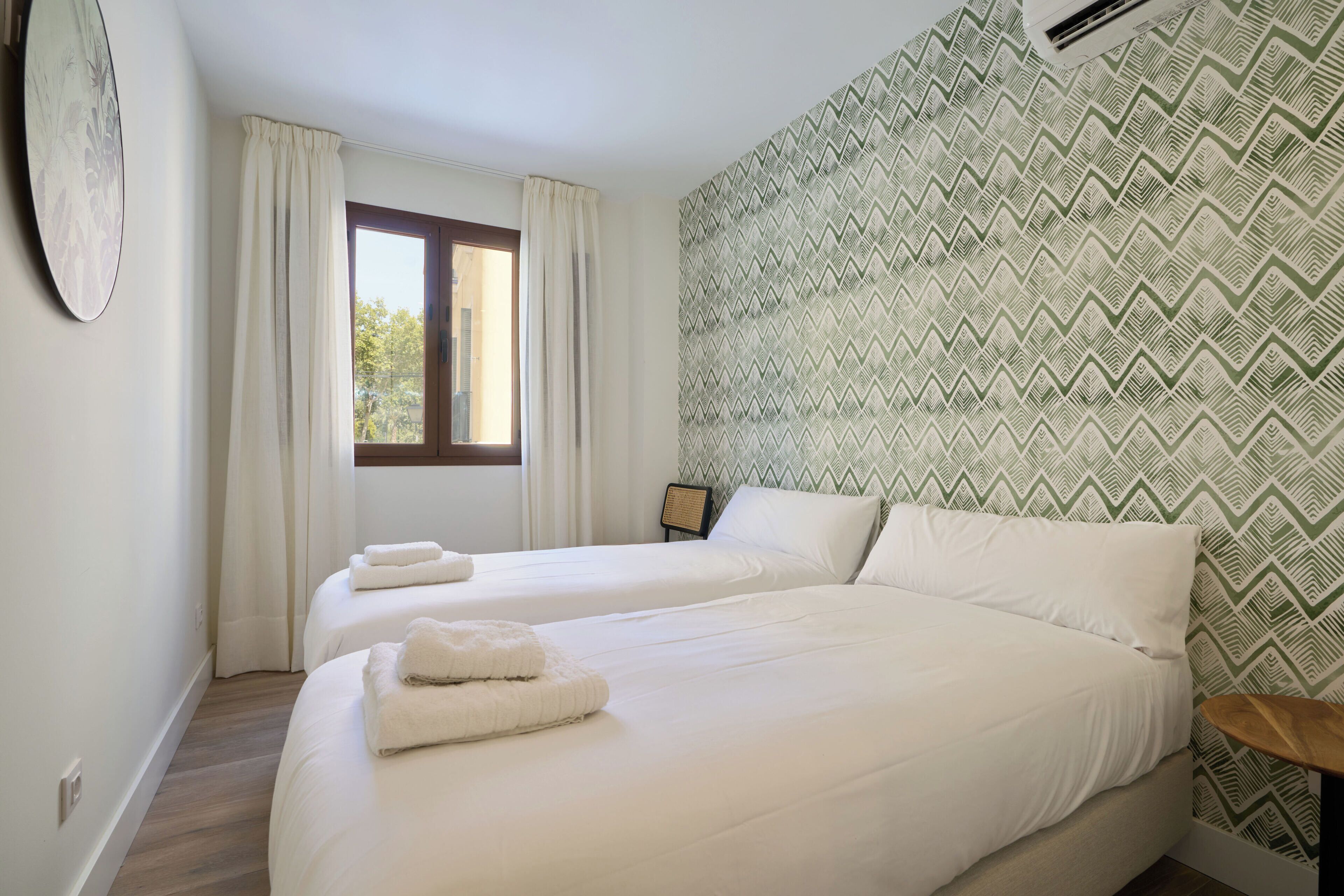 A hotel room with two beds, a window with curtains, a patterned wall, and a clock on the wall.