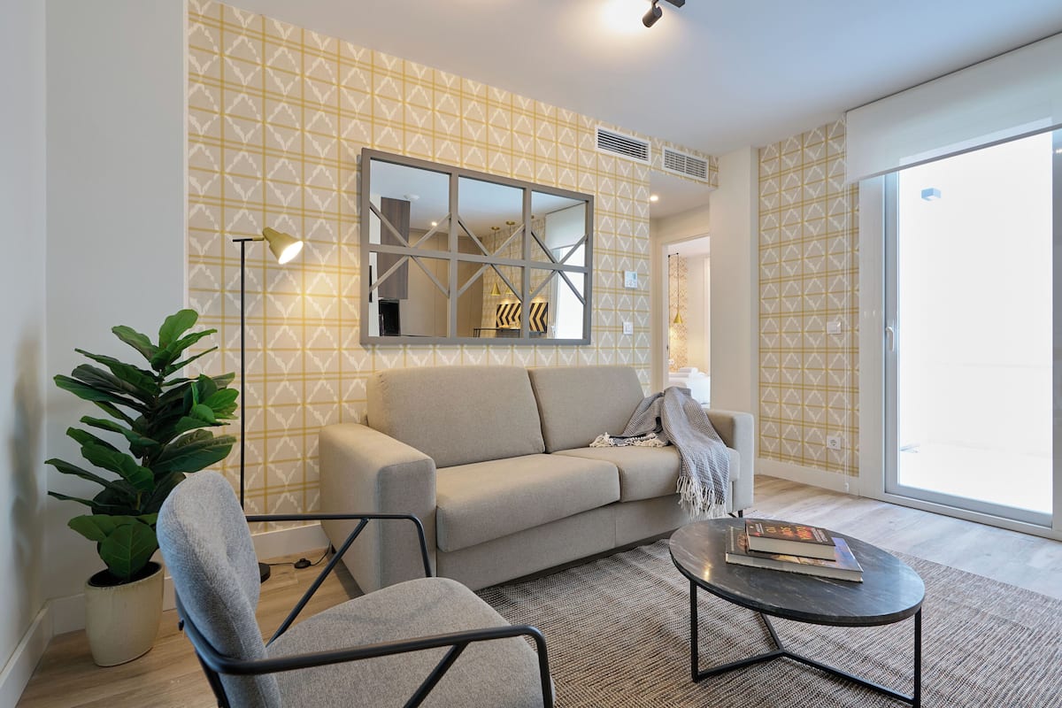 Un salon moderne avec un canapé, un fauteuil et une table basse. Un grand miroir est accroché au mur, et une plante en pot est posée à proximité.