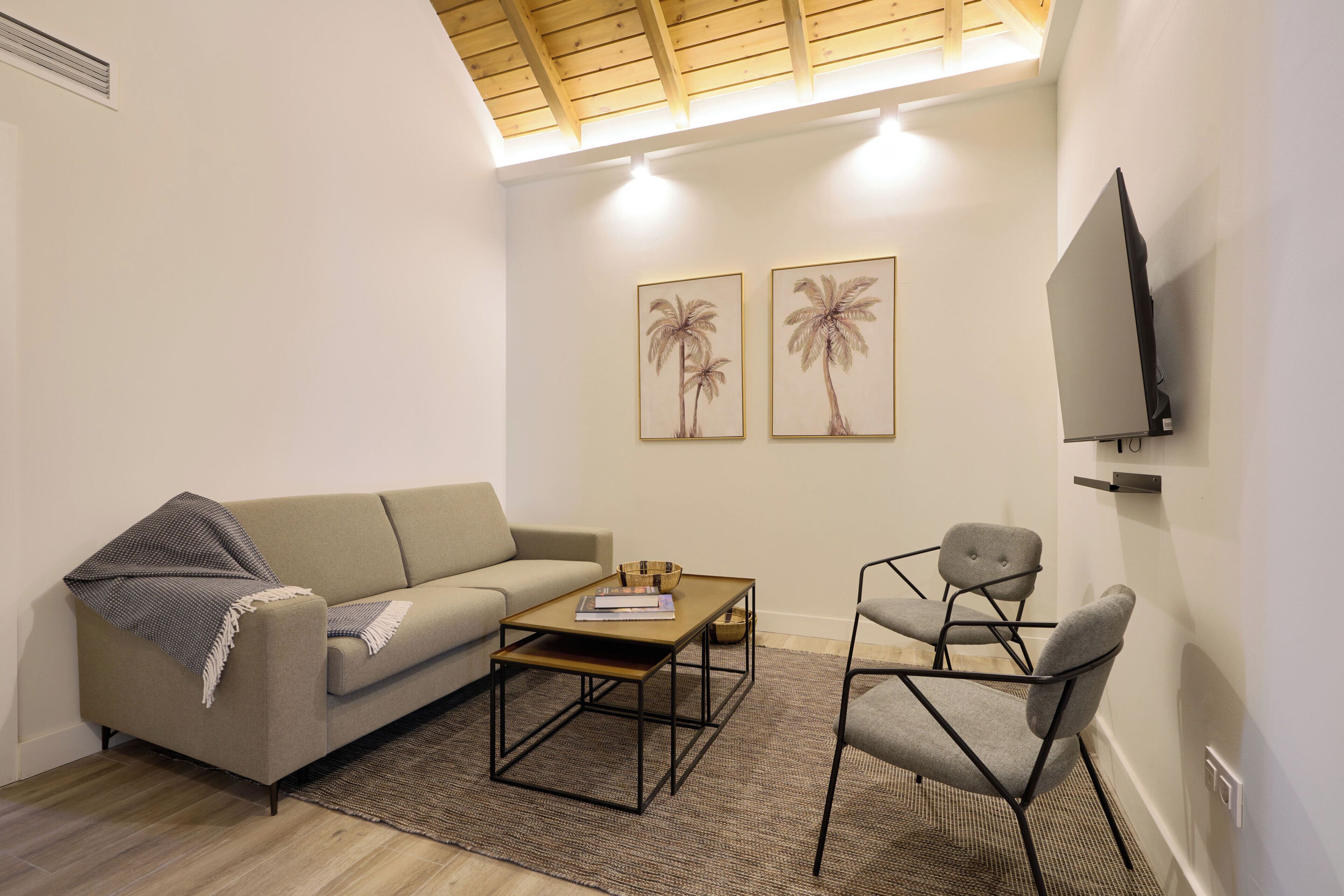 A living room with a sofa, coffee table, and chairs, a flat-screen TV mounted on the wall, and two framed pictures of palm trees.