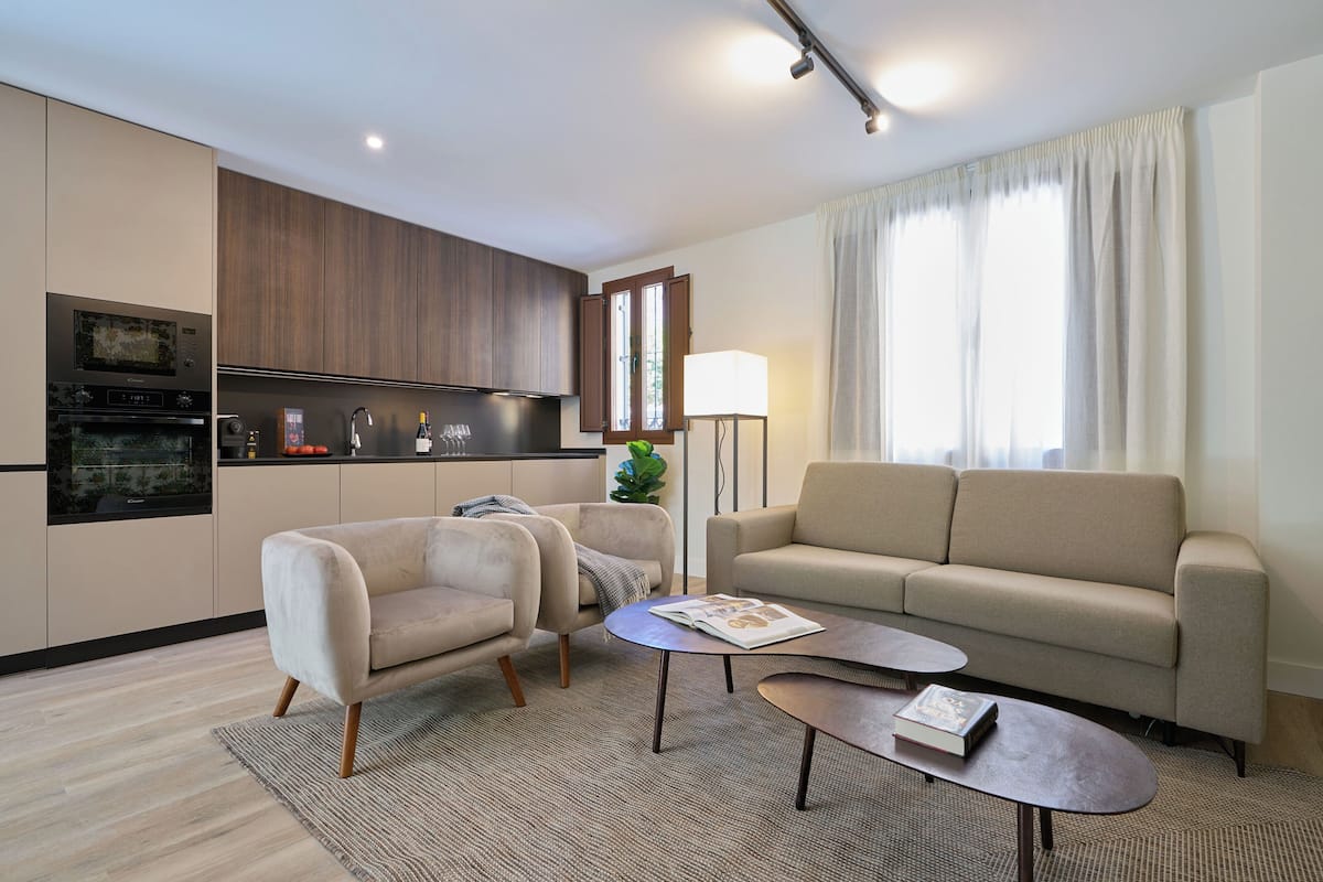 Un salon moderne comprenant un canapé, deux fauteuils, une table basse et un coin cuisine équipé d'appareils électroménagers.