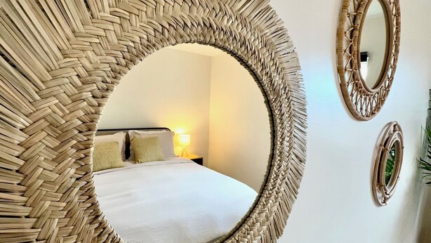 A room with a large woven mirror on the wall, a smaller mirror, and a bed with a white comforter. - Casa Fifi Coral (Miami Beach)