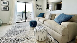 A modern living room with a beige sofa, blue and white cushions, a patterned rug, and a glass door leading to an outdoor balcony.
